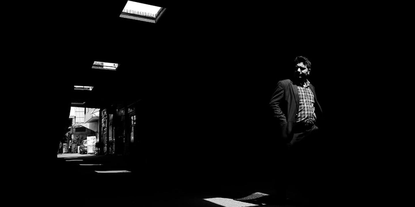 A man walks through a dark alley, looking to one side, illuminated only by roof lights.