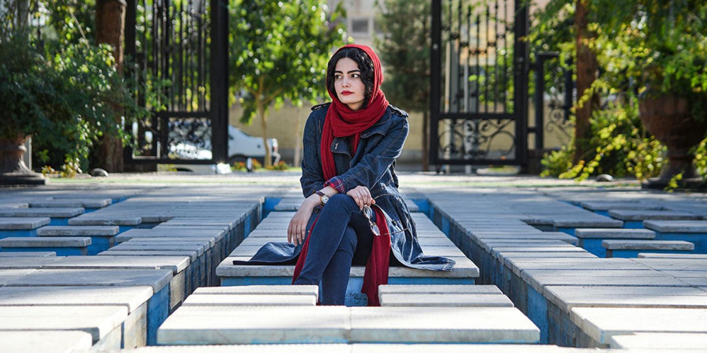 A woman wearing a headscarf sits, looking pensive, amid a grid of concrete seats.