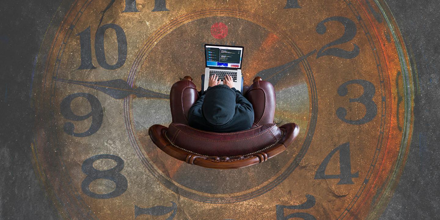 Looking straight down on someone sitting in an armchair working on a laptop. They are surrounded by clock numerals and hands on the floor.