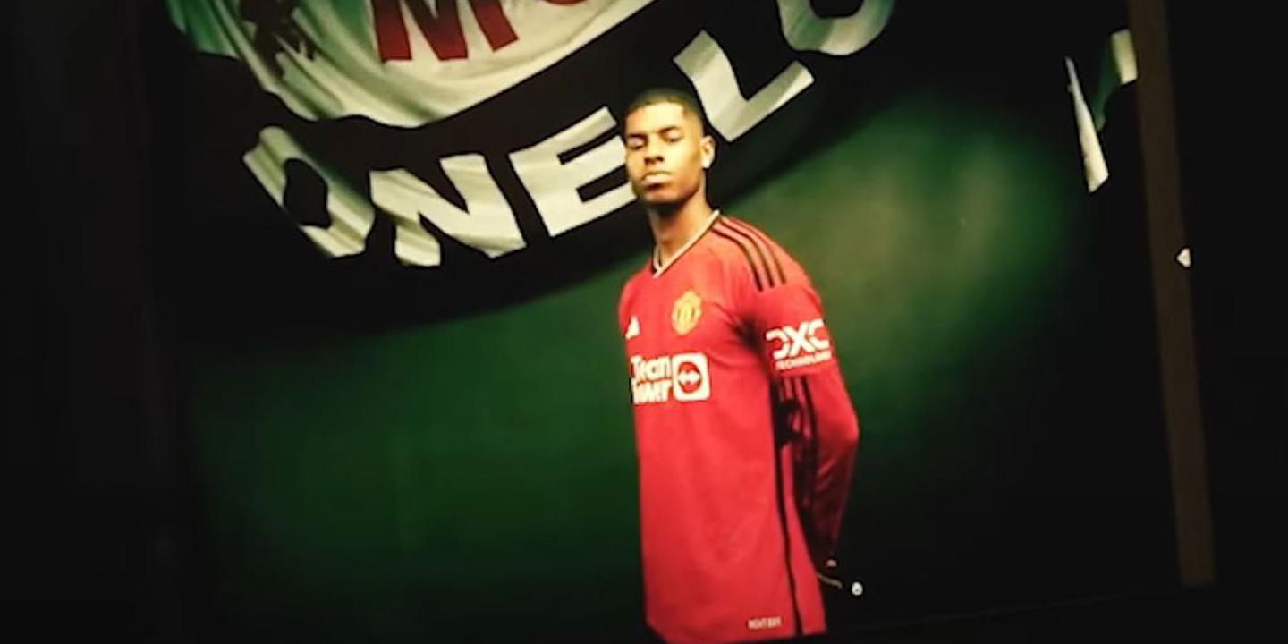 A professional footballer wearign a red top stands proudly beneath a white and black banner.