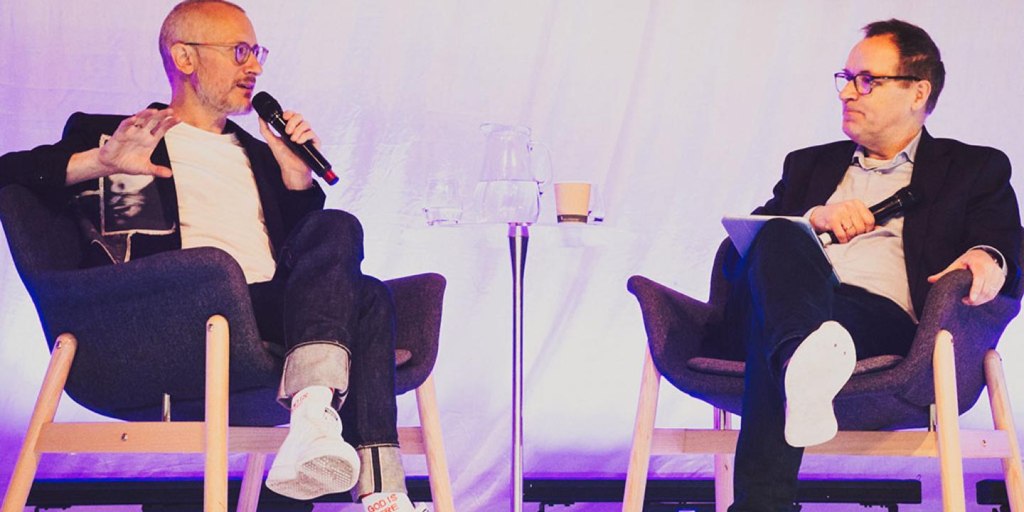 Two people sit on a stage in a relaxed manner, having a converation.