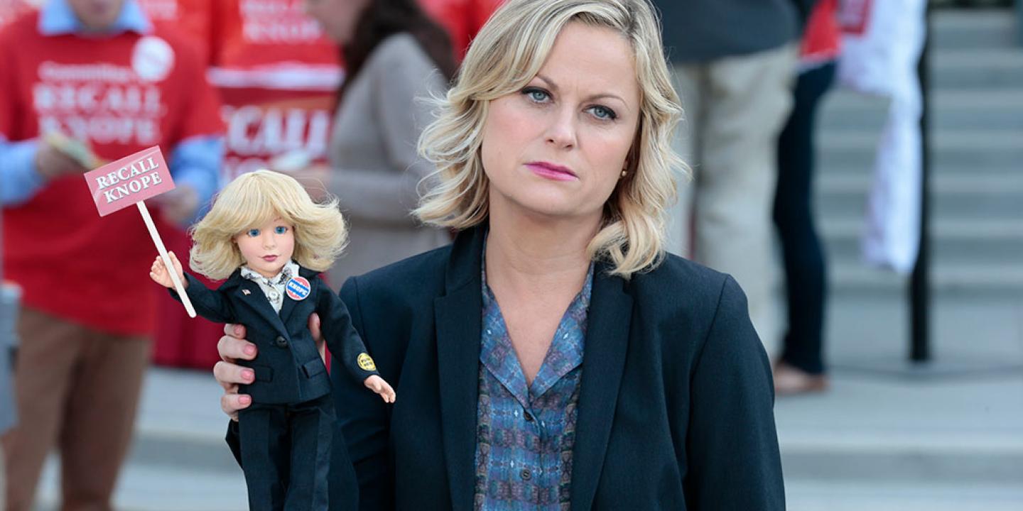 A deflated looking woman stands aside from a protest rally, holding a small doll of herself that reads Recall Knope'.