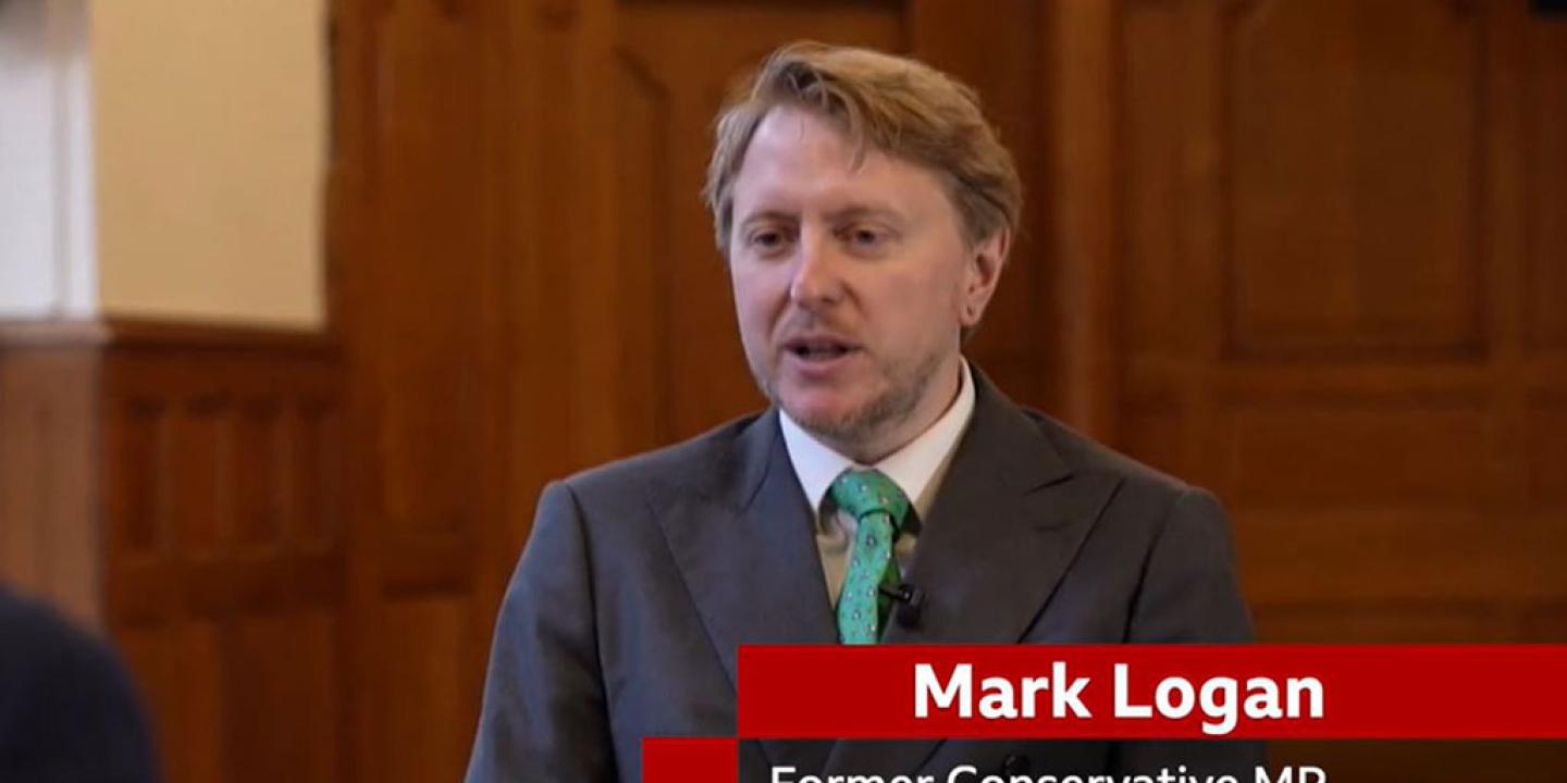 A man sits in a TV interview, below him is a captions stating 'Mark Lognan. Former Conservative MP