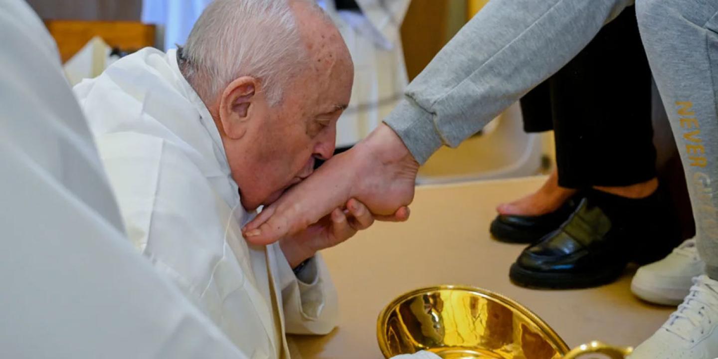 The Pope, wearing white, kneels, crades a bare foot, and kisses it.