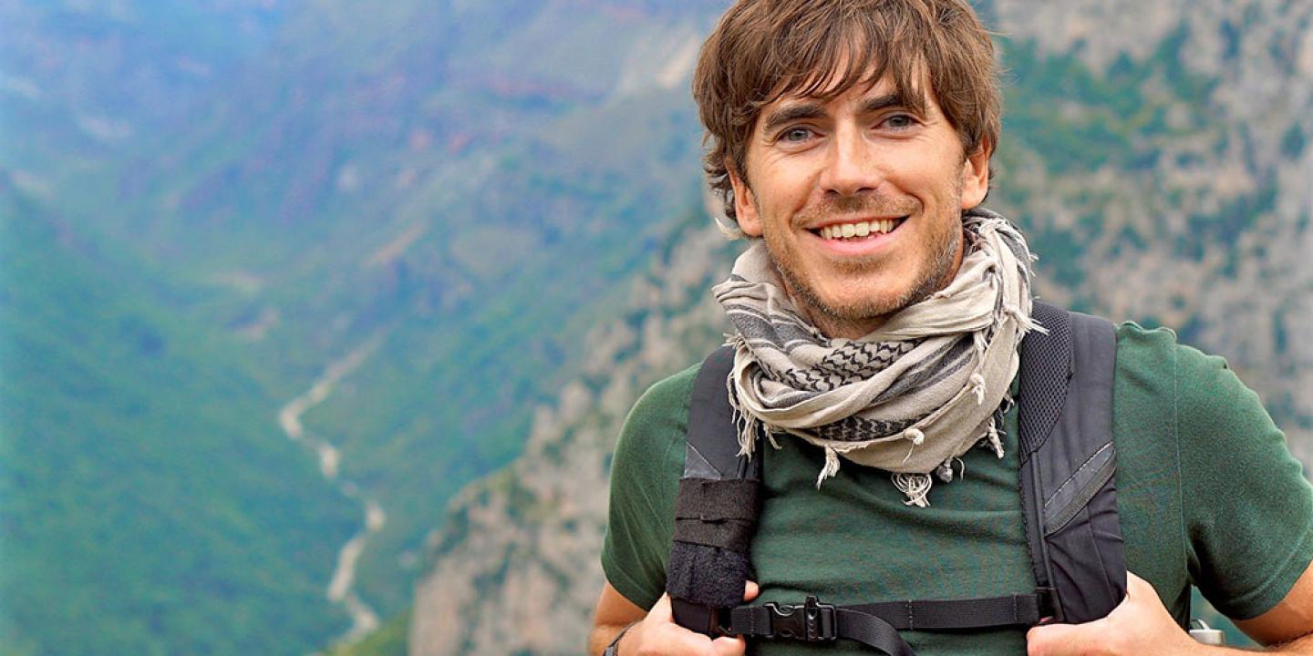 An enthusiastic hiker stands in front of a view down a valley, smiling and holding his backpack straps.