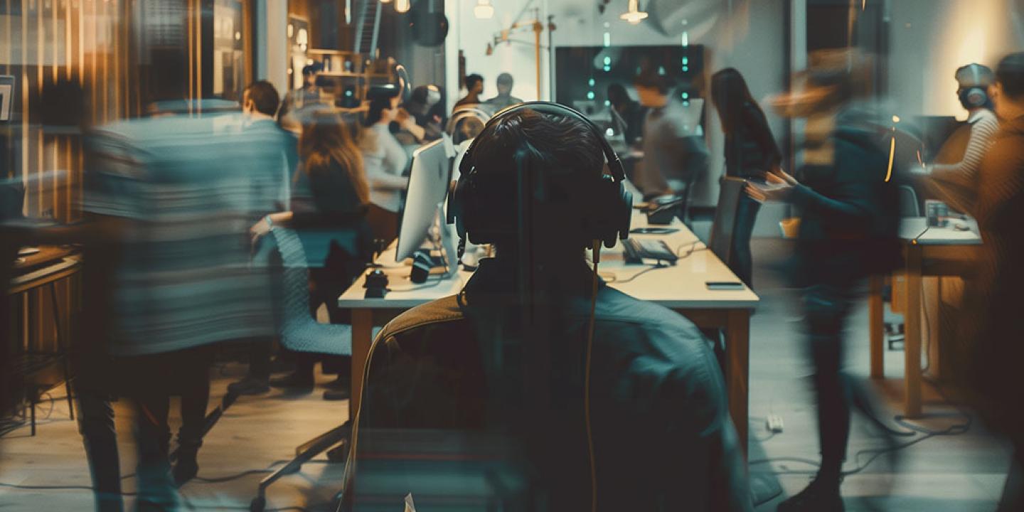 A office worker wearing headphones looks out of a hectic and loud office space around which people are moving