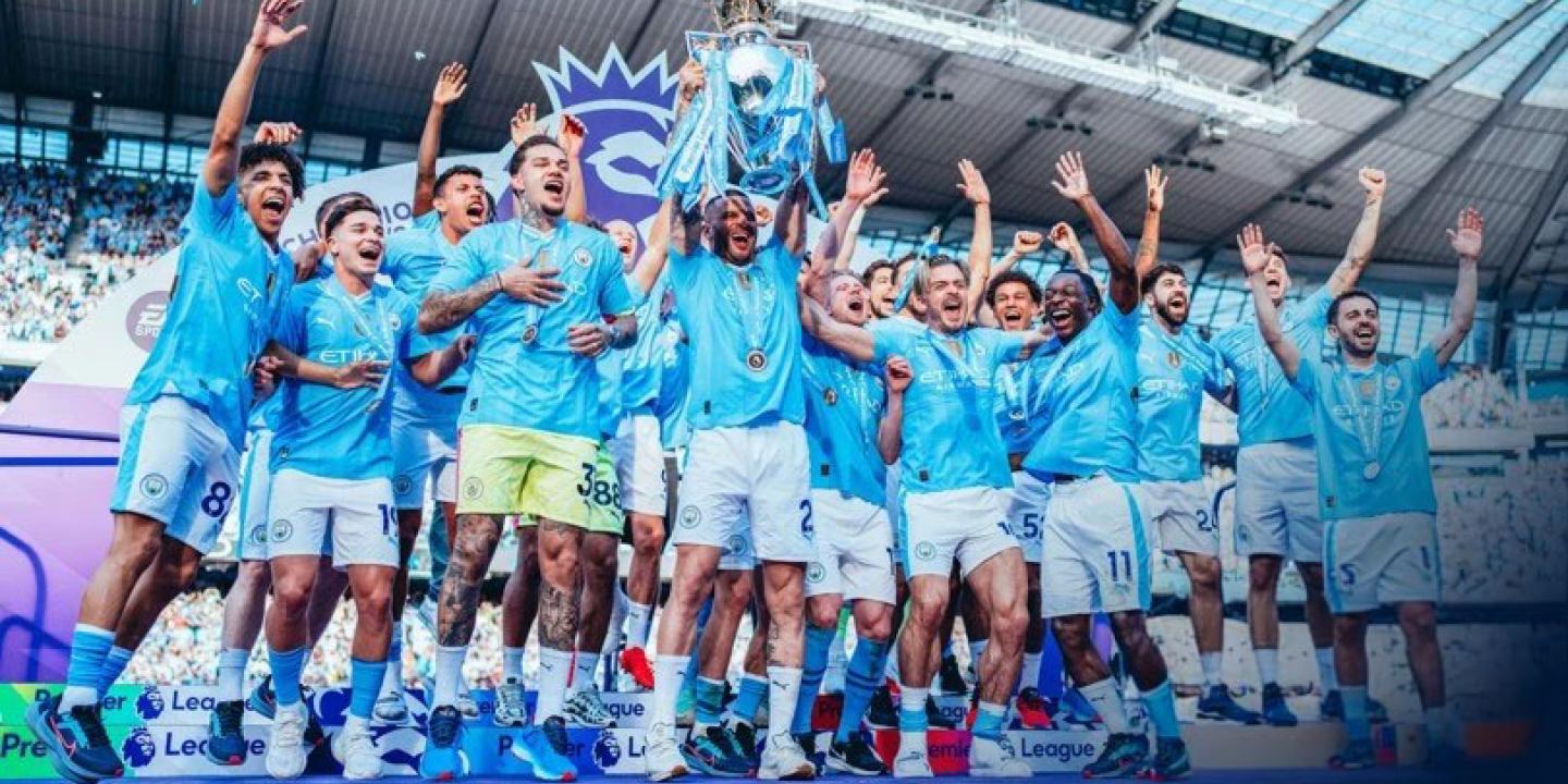 A football team wearing a sky blue kit leaps for joy holding a trophy.