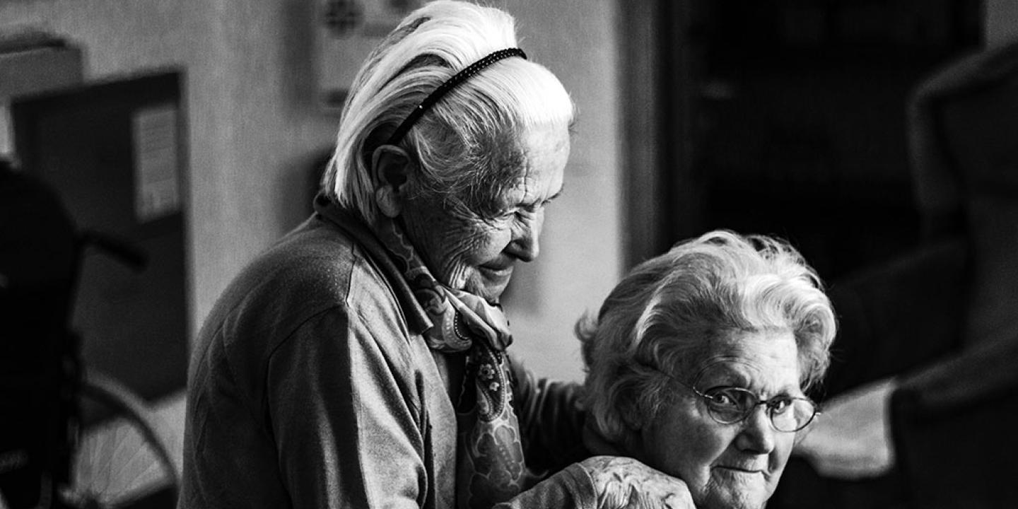 A elderly women in a care home stands and places her hands on the shoulders of a seated woman.