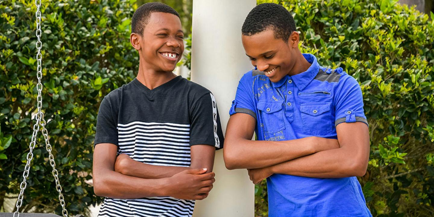 Two teenager lean against a rail, arms crossed, and laugh together.