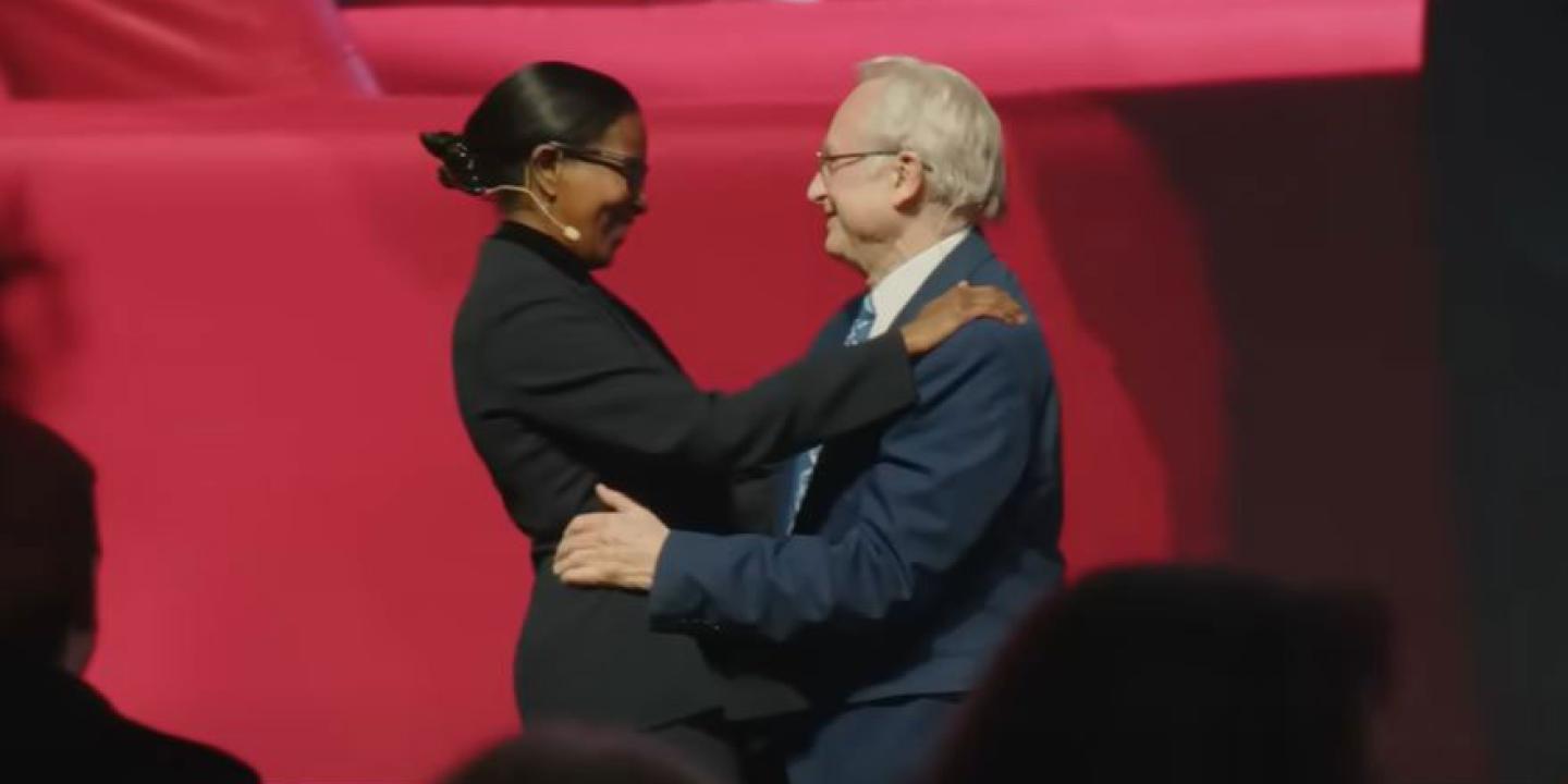 A man and woman speaker on a stage greet and embrace each other.