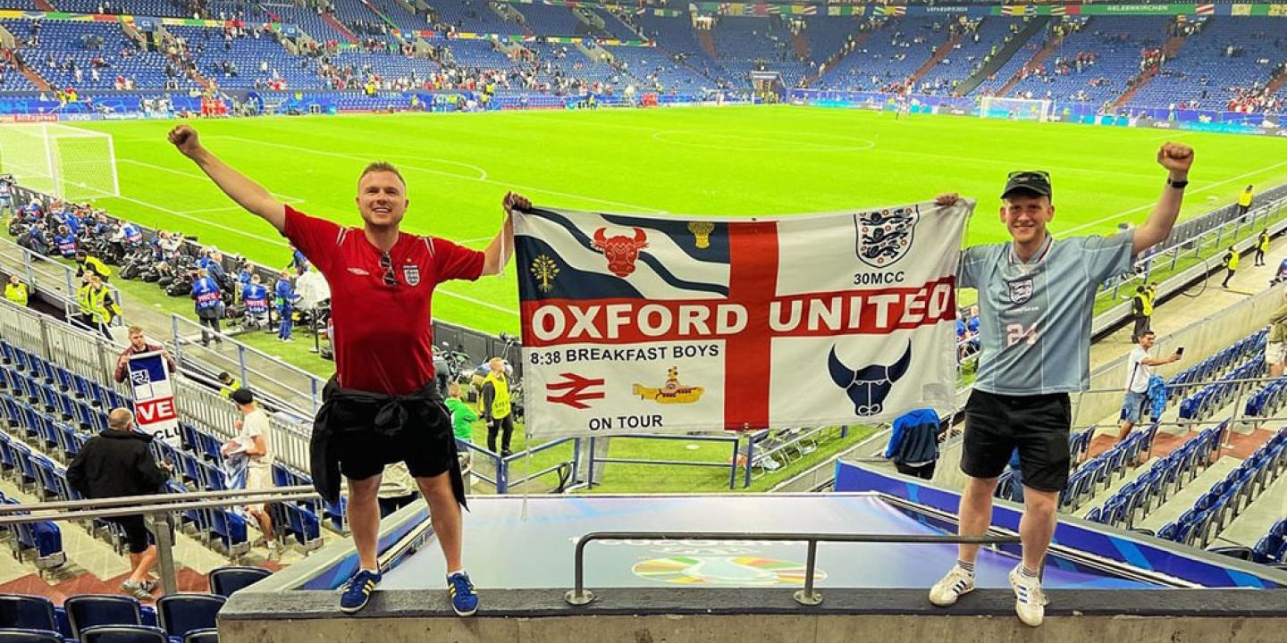 Two England fans stand in stadium seeting holding up their flag.