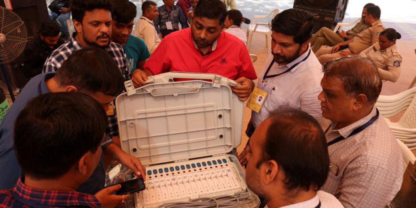 official surround an open case containing an electronic voting terminal and cables.