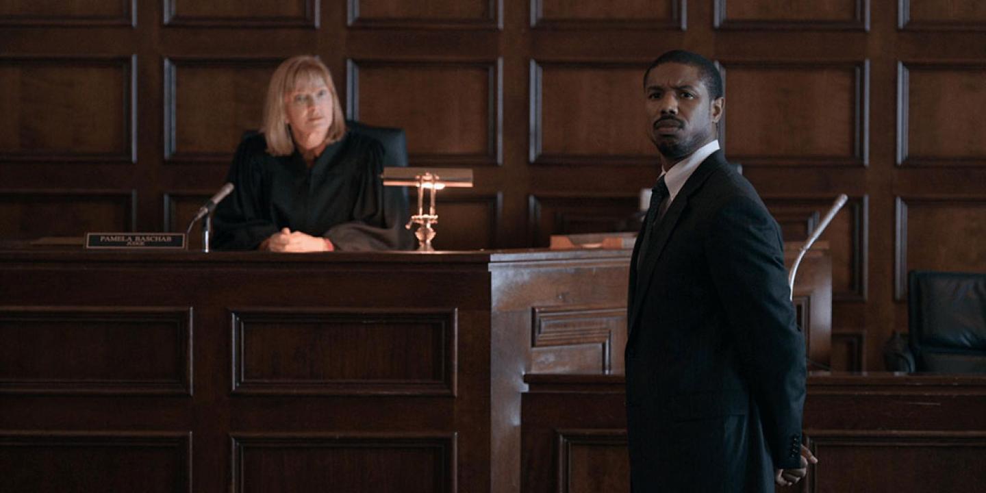 In a court room a judge looks out across it as a lawyer standing addressing her turns his head to look.