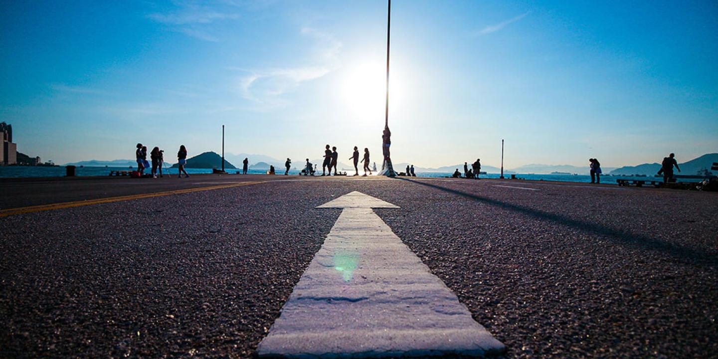 A white arrow on tarmac points towards a setting sun and people walking by a silhouetted lamp post.