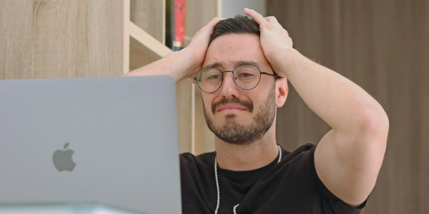 A man staring at a laptop grimmaces and holds his hands to his head.