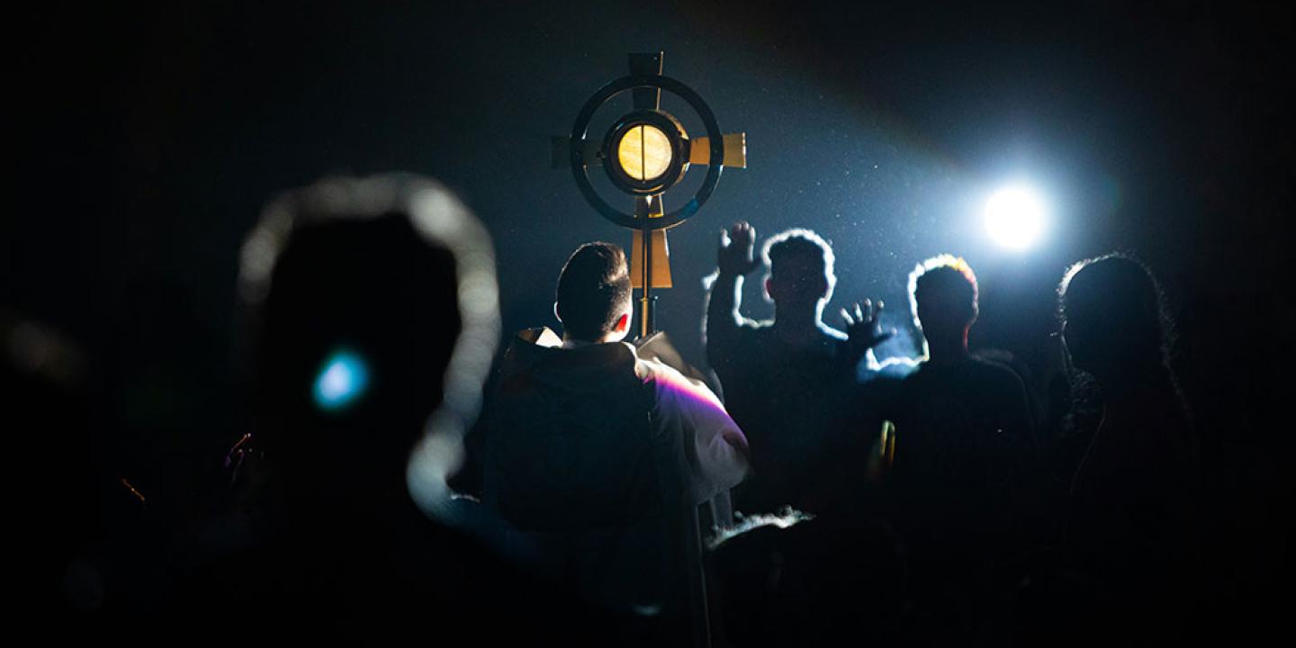 A cross held aloft is illumminated by a shaft of light that also reveals hands raised in priase.