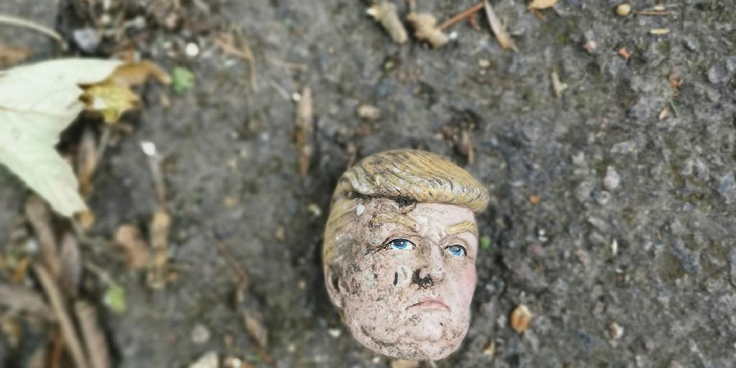 A severed doll head, resembling Donald Trump, lies on dirty ground.