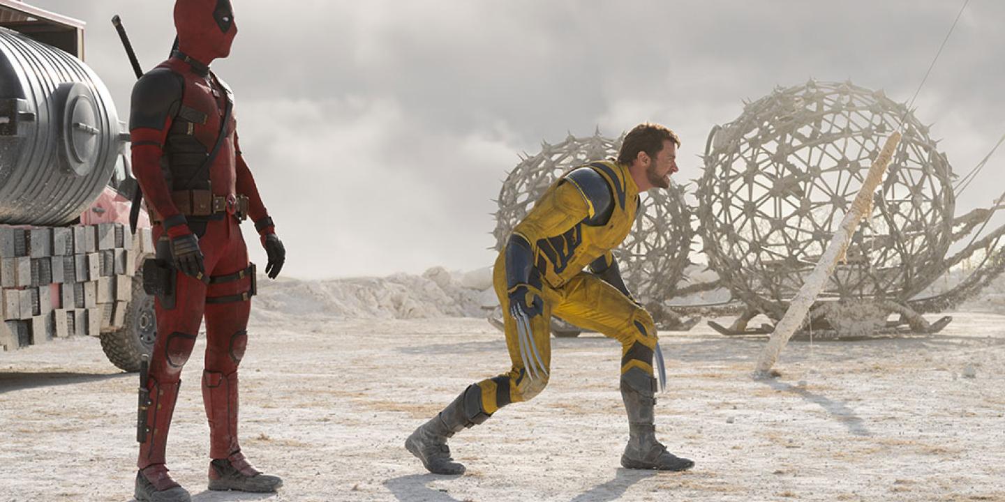 Two superheroes, deadpool and Wolverine, stand and crouch respectively, in a desert like place.