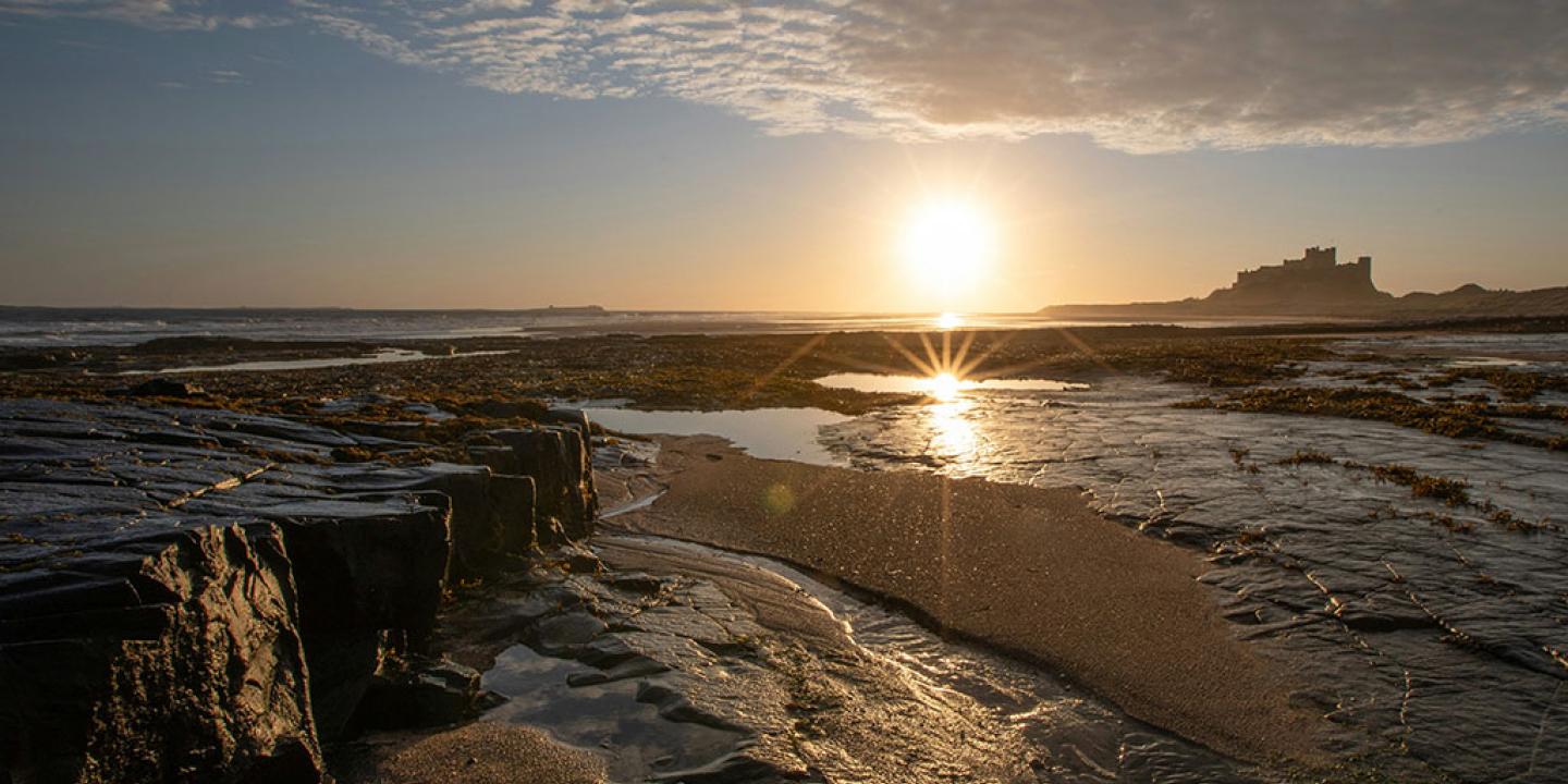 A rockpool on a beach reflects the sun, a castle stands beyond the sand dunes