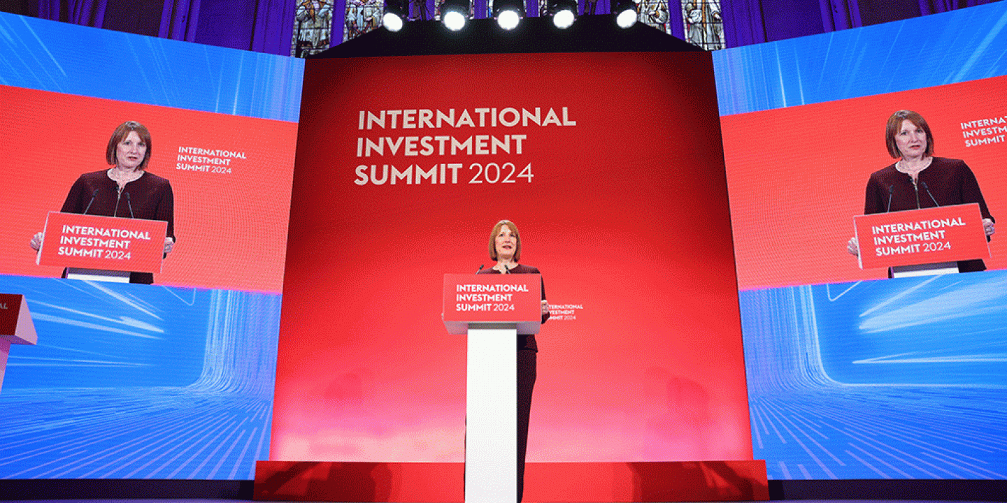 A woman stands behind a lectern against a blue and red backdrop.