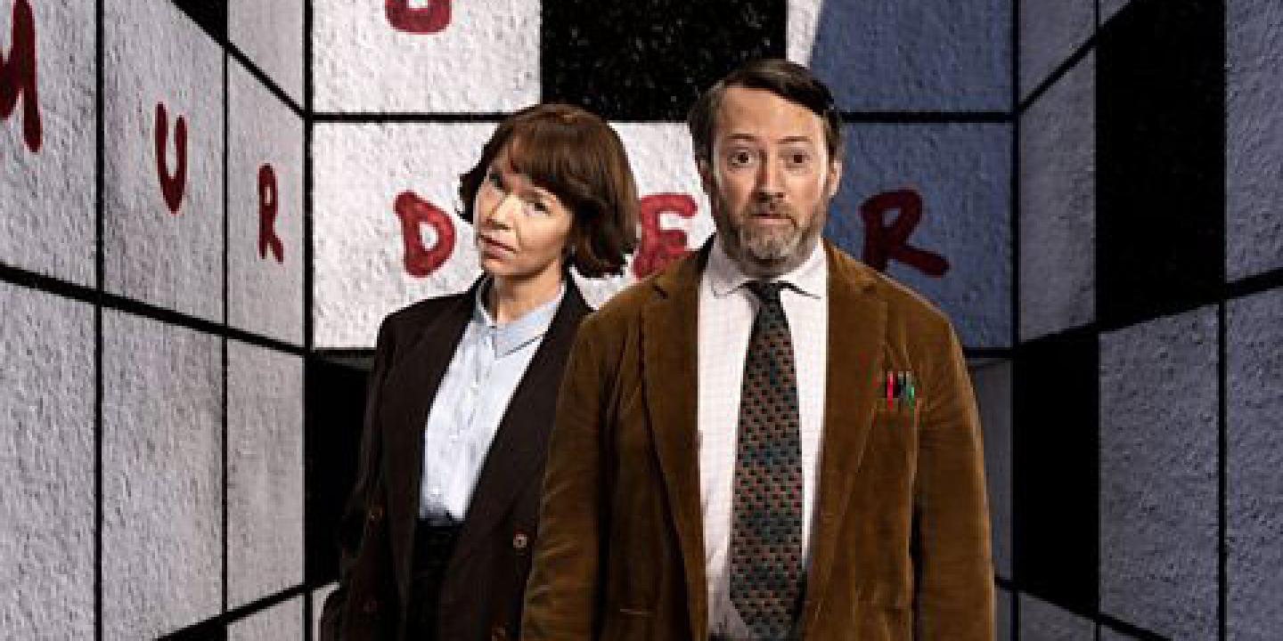 Two TV characters, a man and a woman, stand in front of a crossword cover walls of a room. 