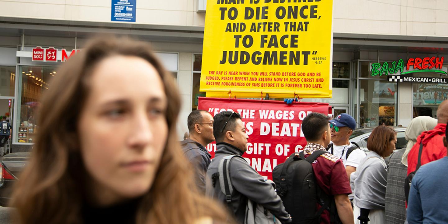 Behind a passer by a street peacher holds up a large yellow sign with a message on it.