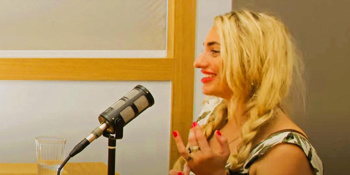  In profile a woman speaks into a microphone and holds her hands up from a desk.