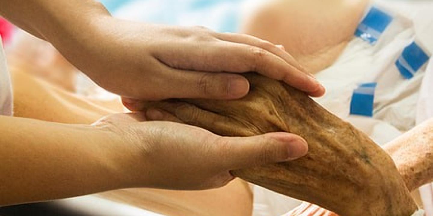 A younger hand holds a wrinkled older hand of someone in a bed.