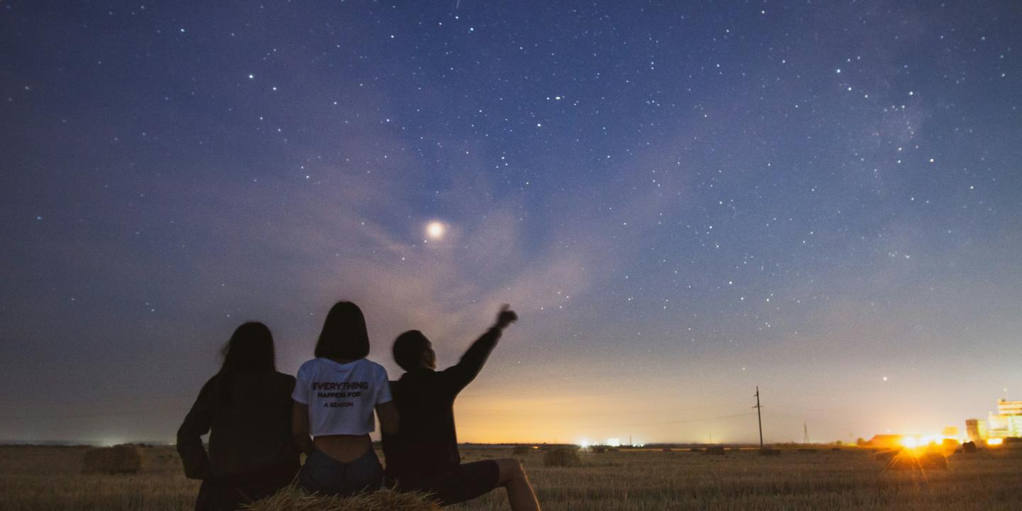 At dusk, three people sit on a field edge and look at the stars emerging.