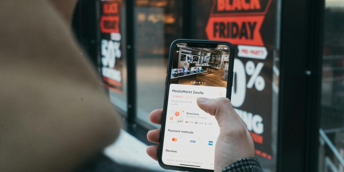 A phone screen is held up, showing a shopping page, behind a window displays a Black Friday Sale banner.