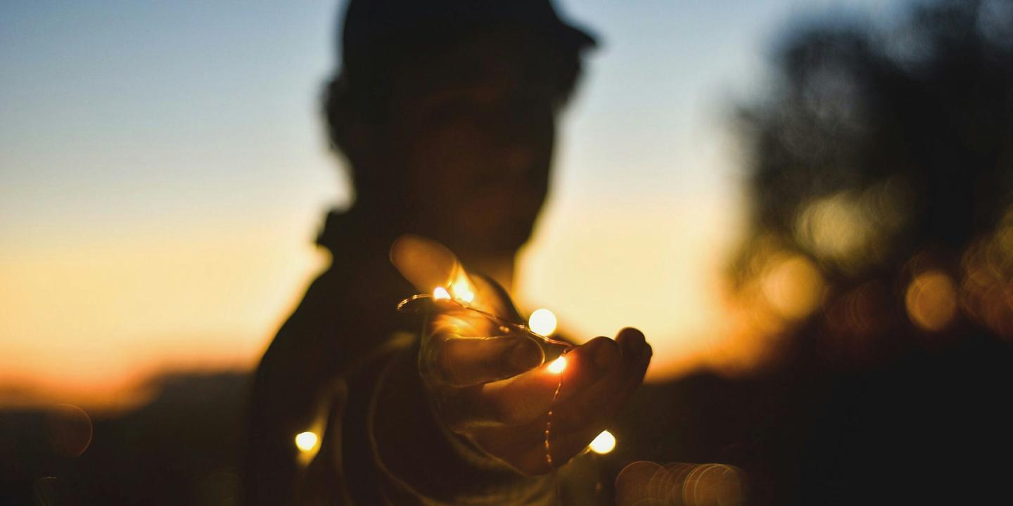 A backlit person at twilight holds a hand out towards the camera, holding some fairly lights