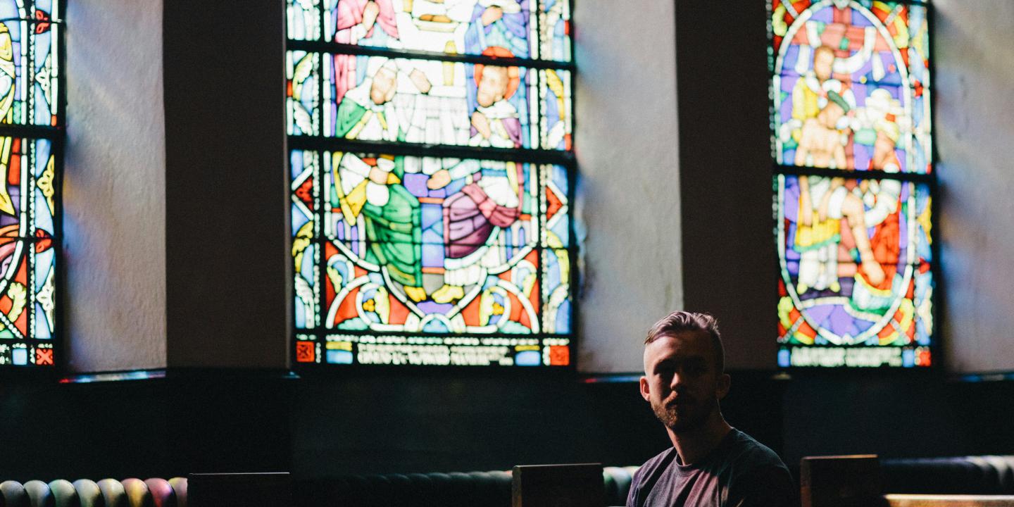 A man sits in a church pew below a colourful stained glass window, looking pensive.