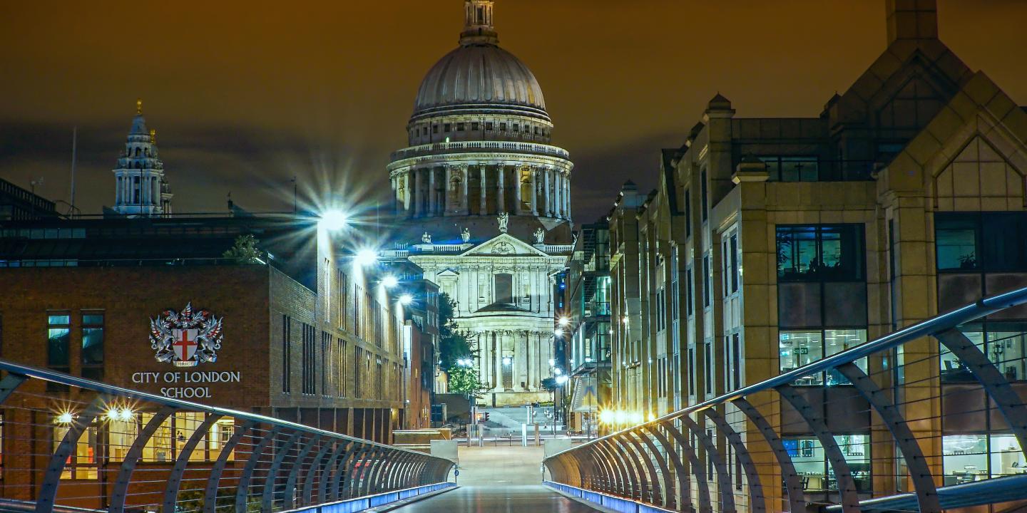 A view from a floodlight footbridge towards a gap between office buildings which reveal a cathedral and its illuminated dome.