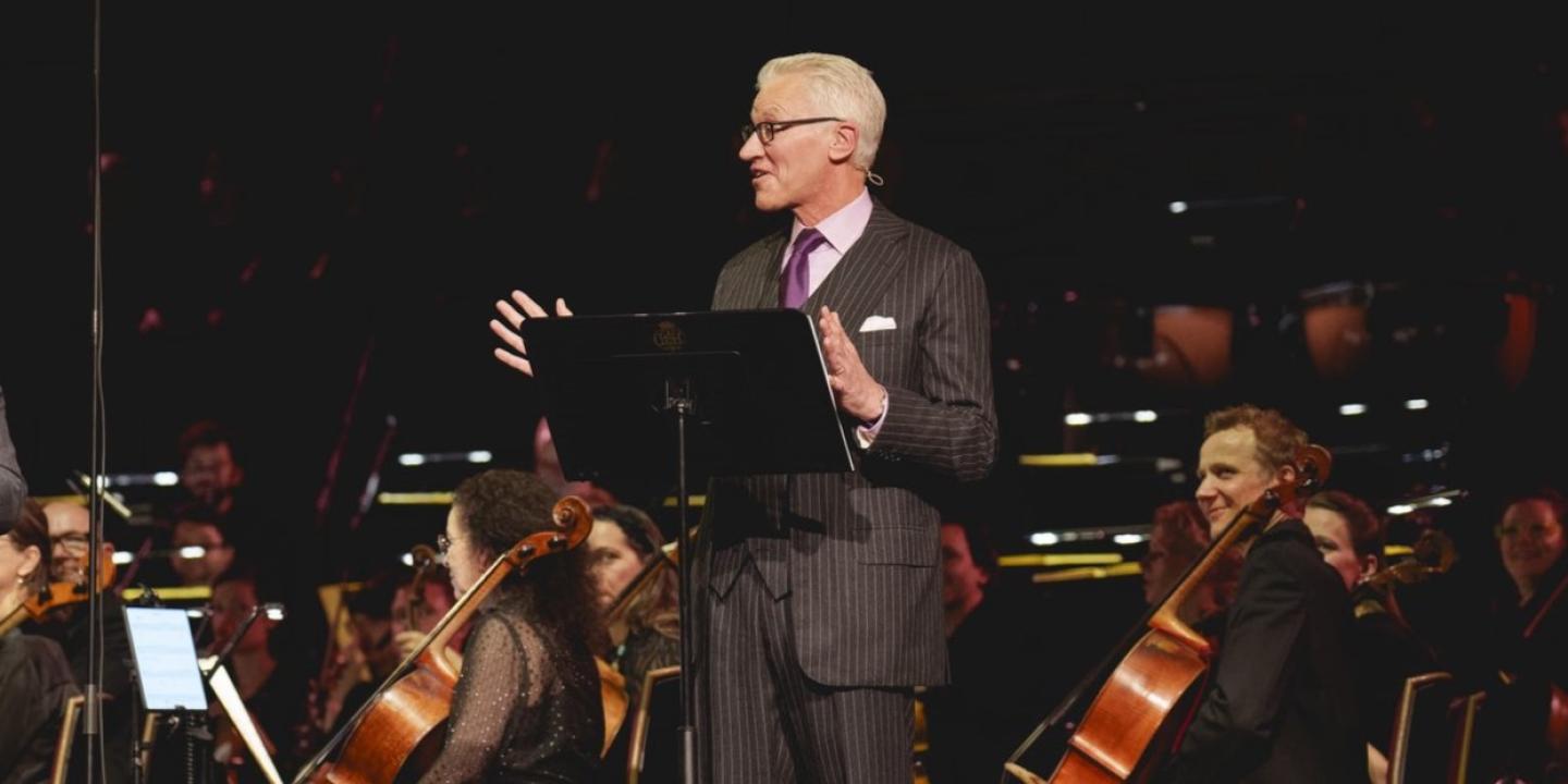 A man in a suit stands in front of a orchestra, by a lectern, gesturing while talking.