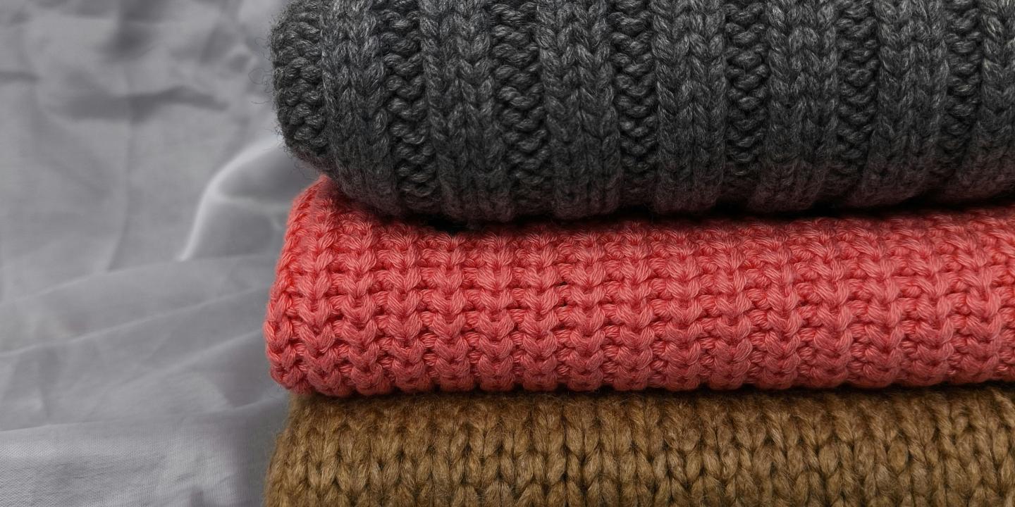 A pile of folded knitted jumpers sit on a grey back ground, one is also grey, the others are red and brown.