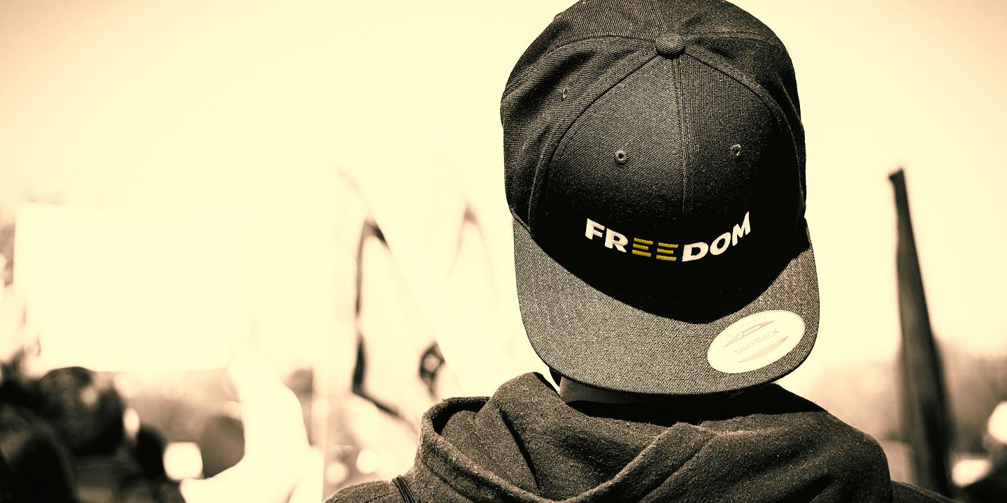 A protester stands with back to the camera, his baseball hat is turned backwards, it reads 'freedom'.