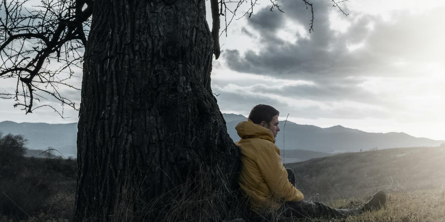 A wrapped-up man sits and leans against a bare tree, as dark clouds give way to sun.