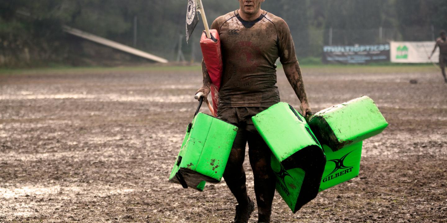 A muddy rugby player carries equipment off a pitch.