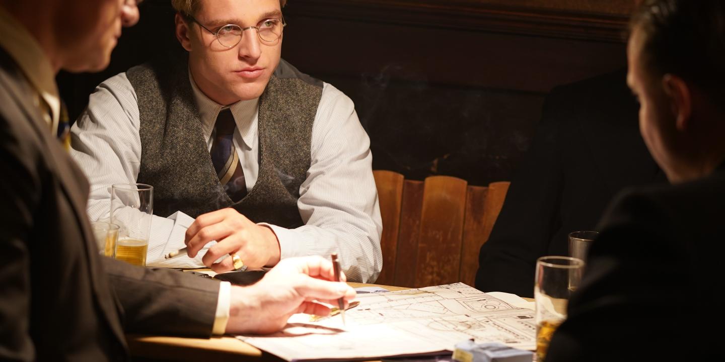 A man dressed in 1930s clothing, sits with others at a table looking pensive.