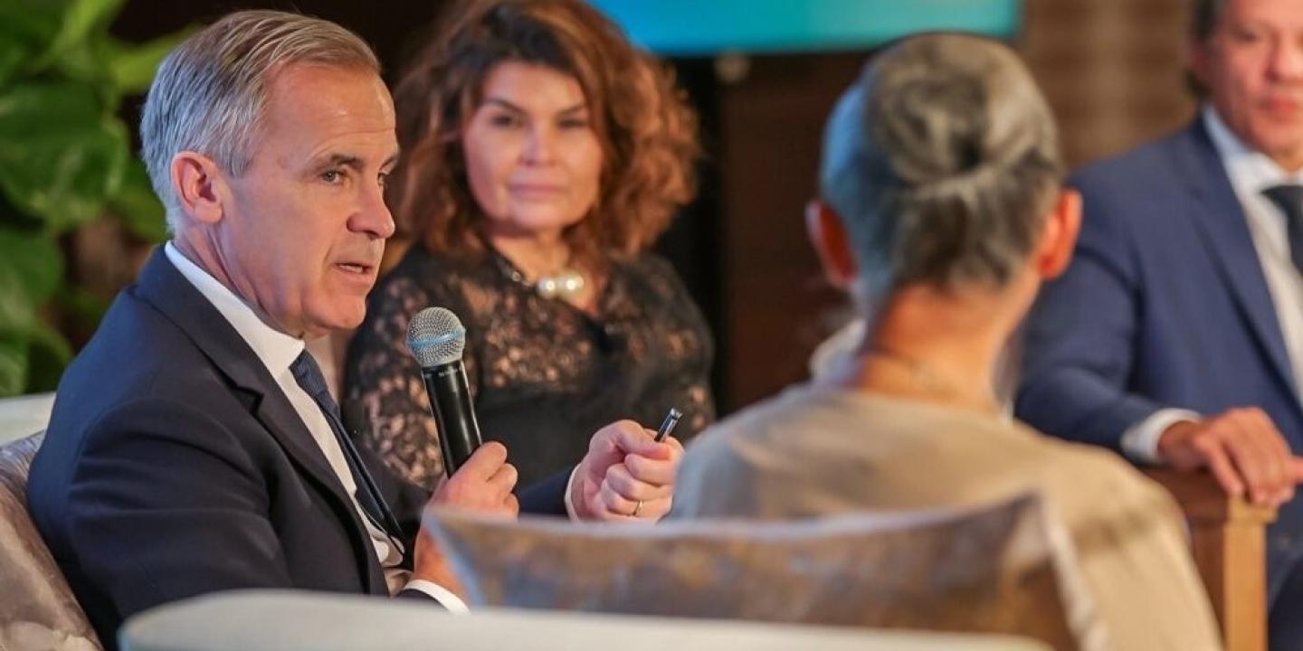 Mark Carney sits between two other speakers, holding a mic.