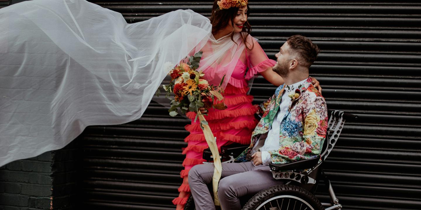 A bride dressed colourfully stands next to her groom, dressed similarly, as he sits in a wheelchair.