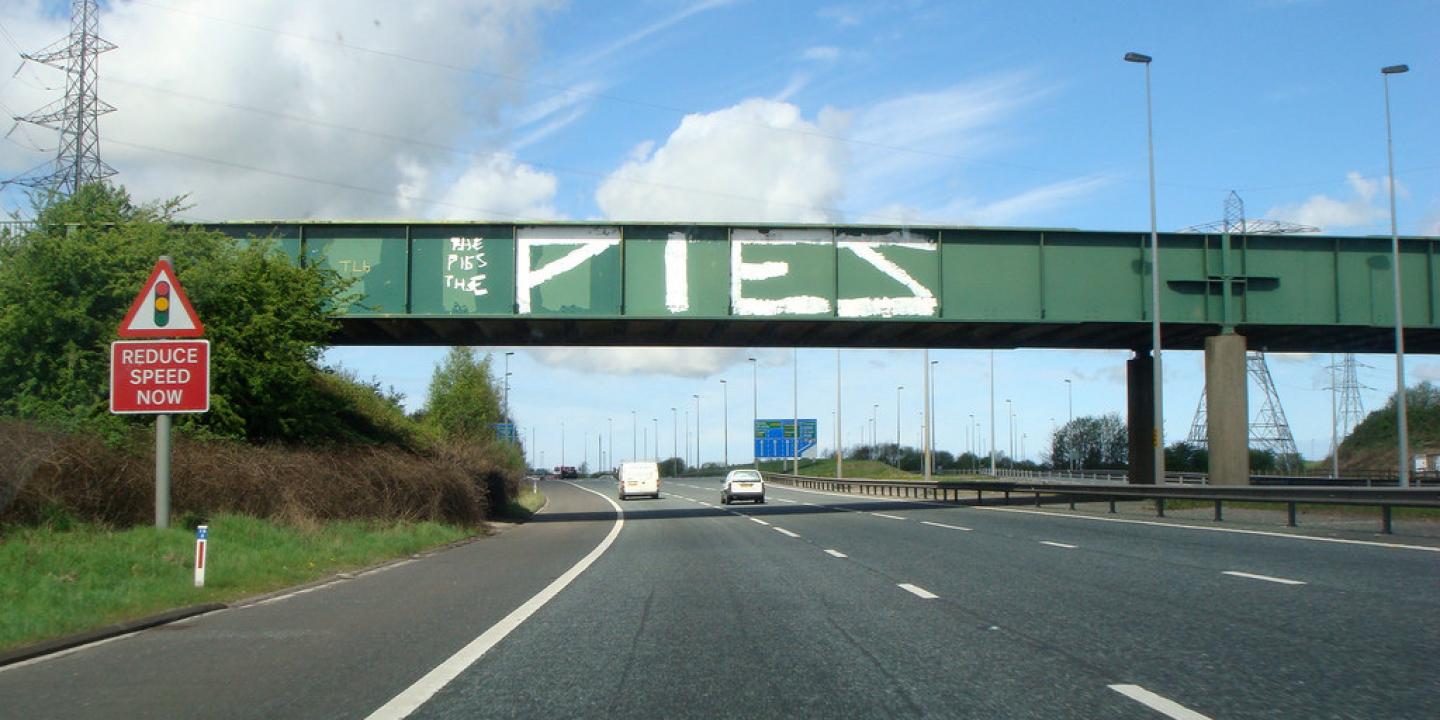 A green bridge spans a motorway, on its side is graffiti that reads 'PIES'