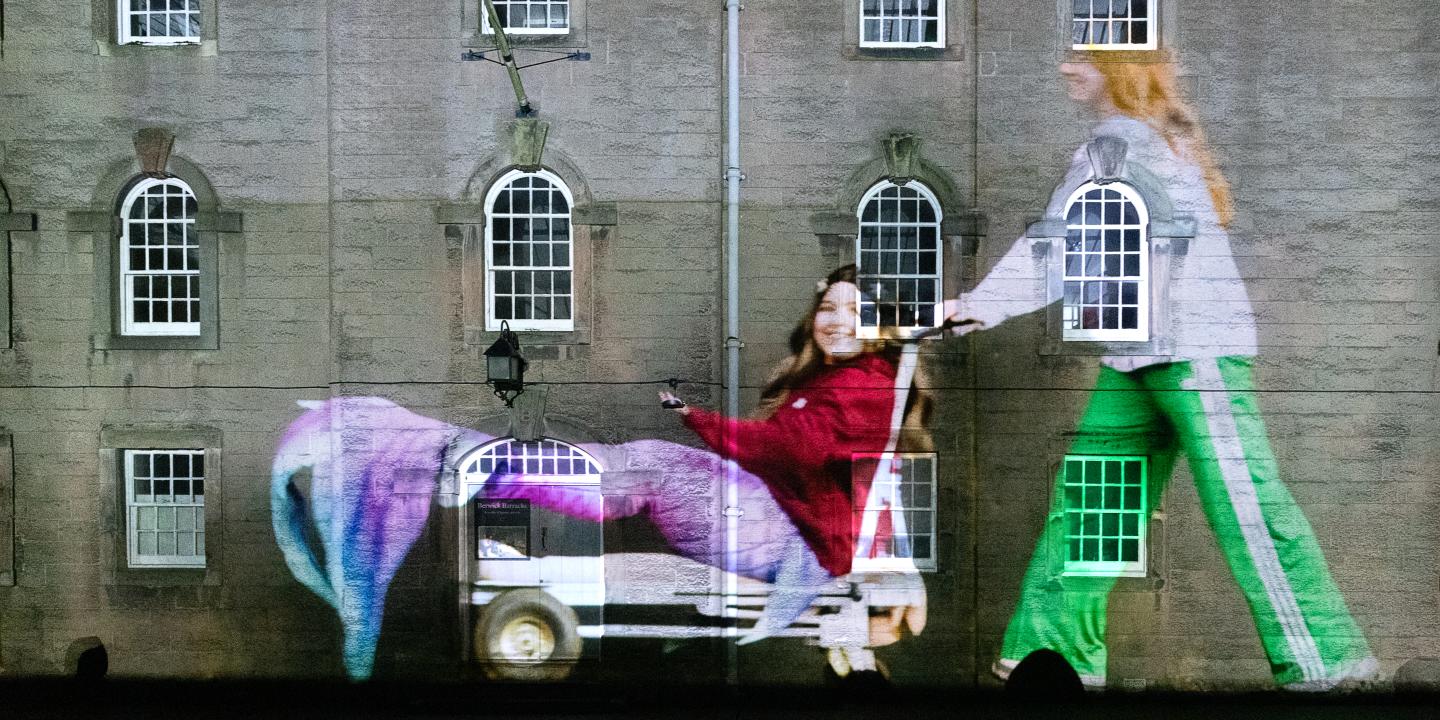 A projected image of a person dressed as a mermaid being pushed on a trolley falls on the wall and windows of an old barracks.