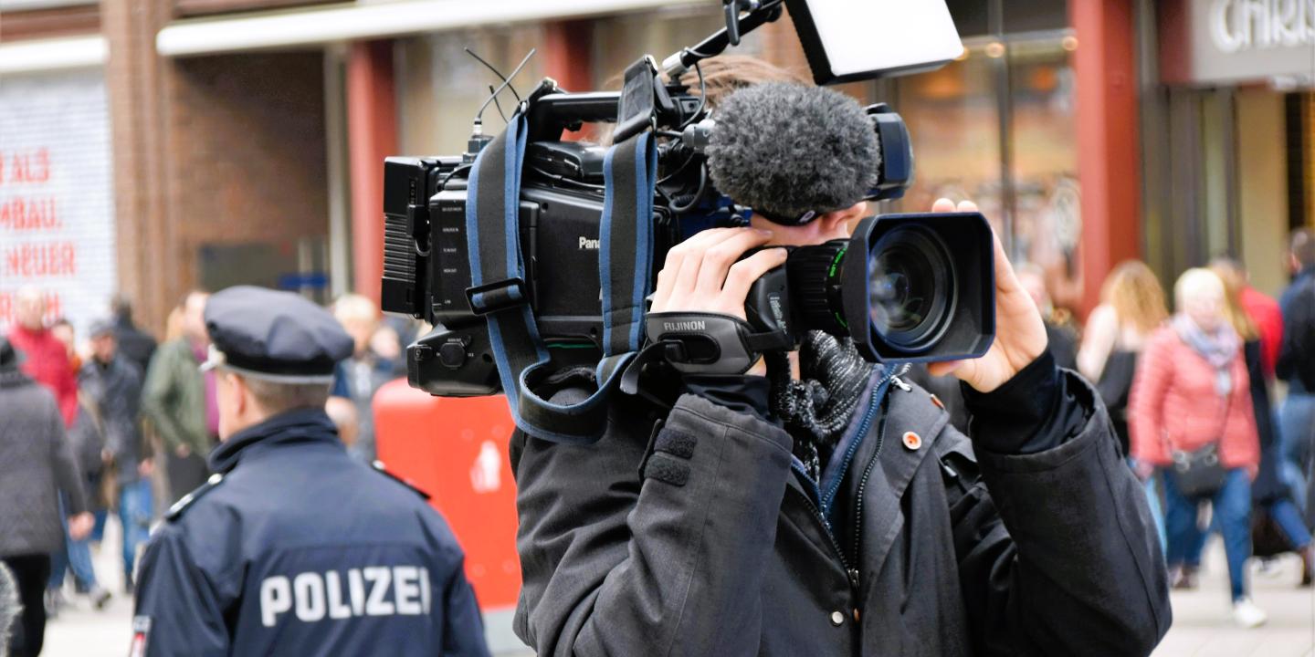 A news cameraman holding a camera, stands back to back to a police officer.