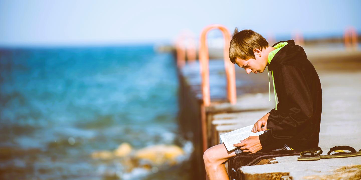 A man sits on a pier intently reading a book on his lap.