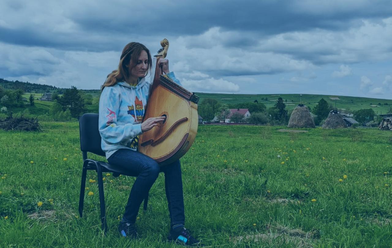 A woman sits on a chair in a field holding a large stringed musical instrument.