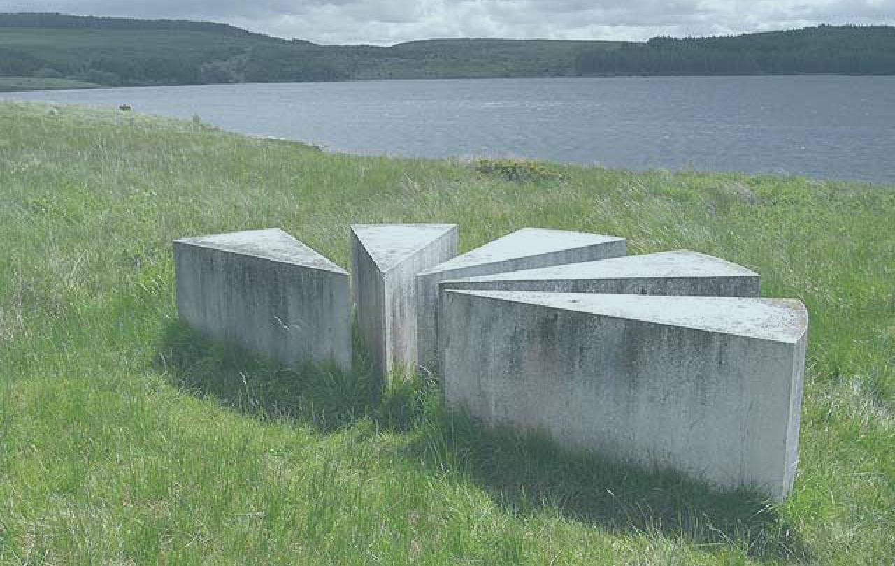 A set of low concrere blocks in the shape of a map's viewpoint symbol sit beside a lake.