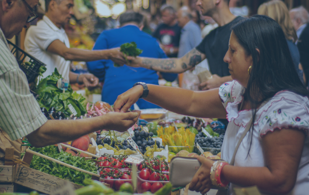 Marketer stall traders and customers reach out arms and hands to exchange fruit and money above piles of produce.