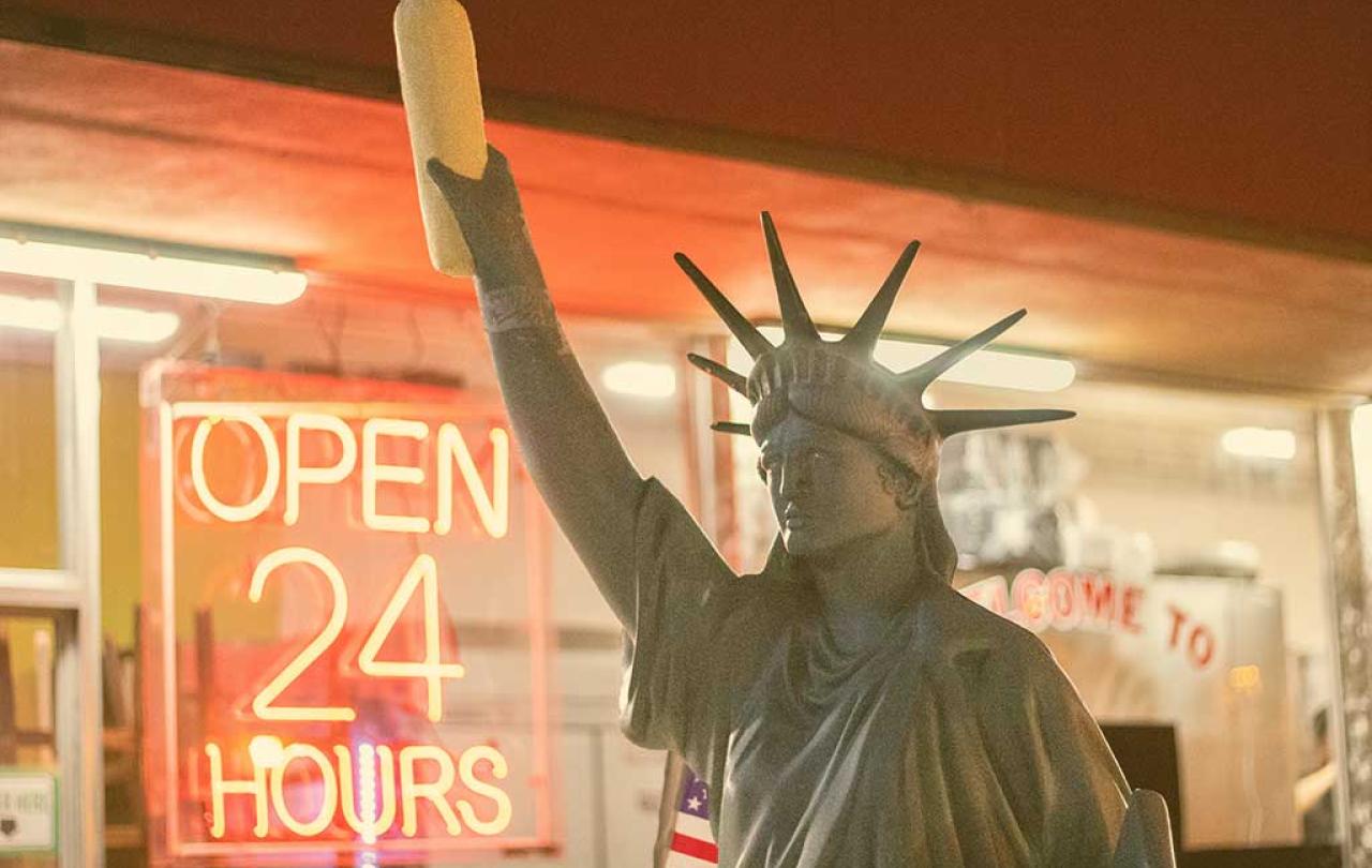 A copy of the Statue of Liberty, holding a stick of bread, stands outside a shop window displaying an 'Open 24 Hours' signs.