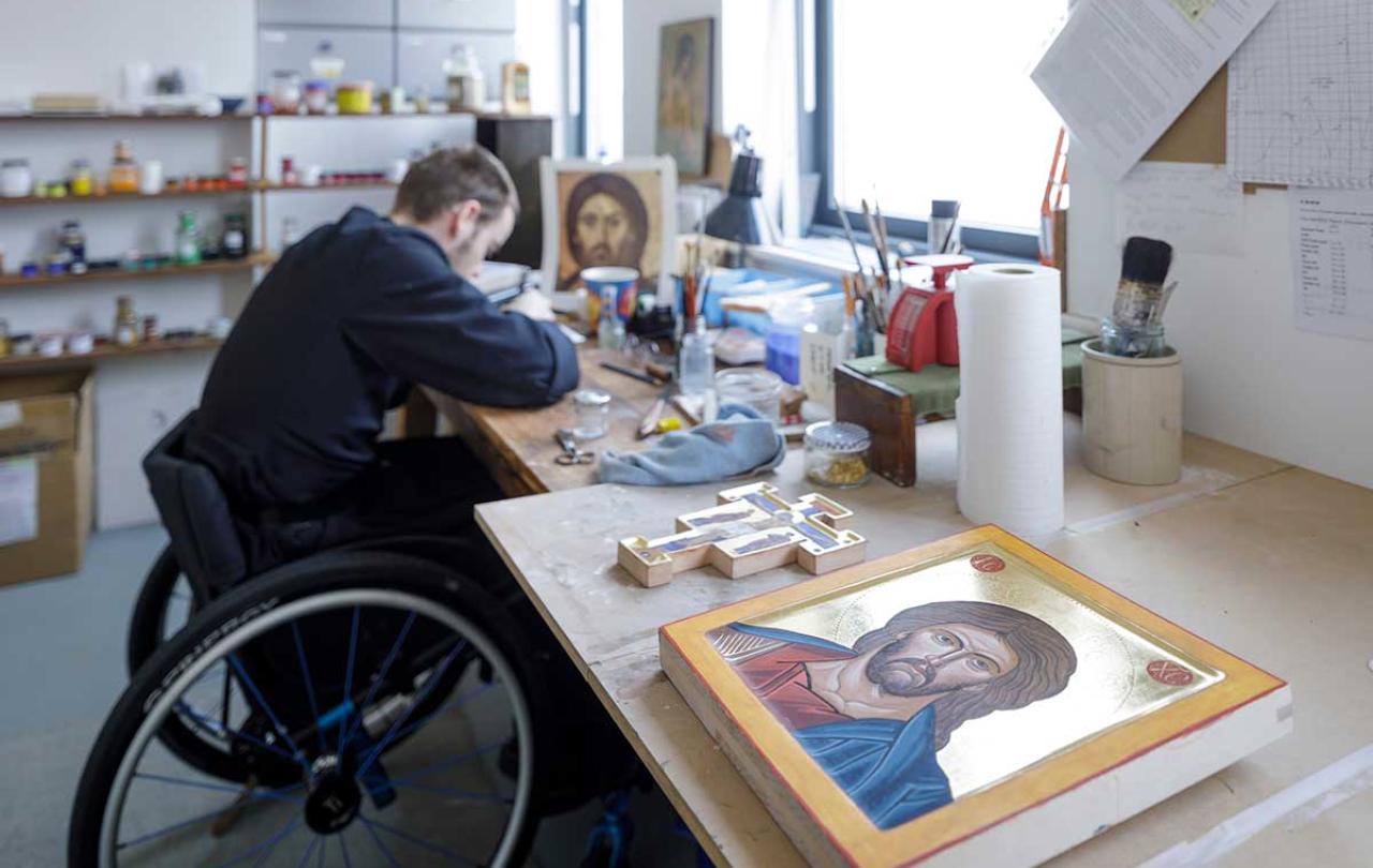 A monk in a wheekchair works on an icon in an art studio. In the foreground is a completed icon. 